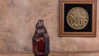 Rustic setting with metal lantern, red-blue glass; framed artwork with Arabic calligraphy.