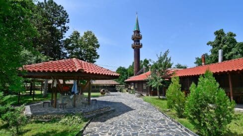 Stone path leads to wooden structure with red roof, tall tower, and greenery in serene setting.