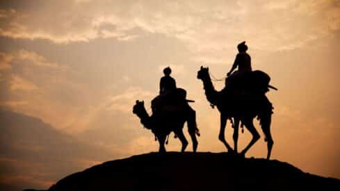 Silhouetted camel riders at sunset on a dune with a cloudy sky.