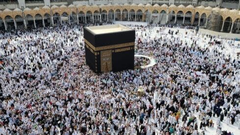 Crowd in white garments around the Kaaba, a black cuboidal structure, in a mosque courtyard.