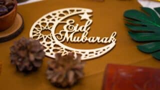 A crescent and a star wooden decoration with "Eid Mubarak" written surrounded by a pine cob and tree leaves.