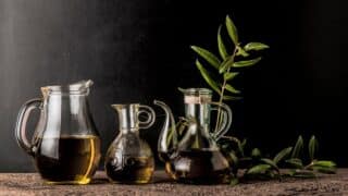 Glass jars of olive oil with branches on textured surface, dark contrasting background.