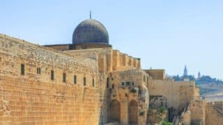 Historical stone building with domed roof, al aqua mosque arched windows, clear sky, and hilly landscape backdrop.