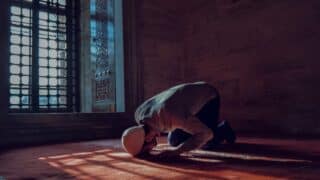 Person kneeling in dim room, praying on carpet; sunlight through lattice window creates shadows.