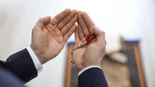 Person in formal attire holds red prayer beads, palms open, indoor setting.