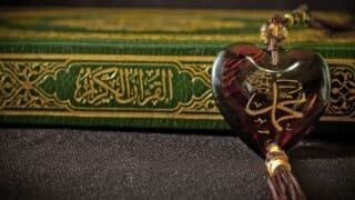 Heart-shaped pendant with Arabic calligraphy in front of a green holy Quran with Arabic script.