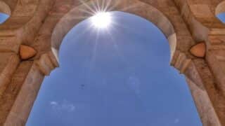 Upward view of stone archway with sunlight starburst and distant bird against blue sky.