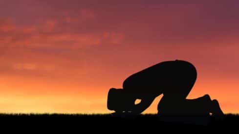 Silhouette kneeling in prayer on grass with sunset sky background.