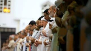 People standing in a line, praying, wearing traditional attire in a well-lit room.