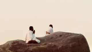 Two people in white cloth sit on a large rock, one praying, against a clear sky.