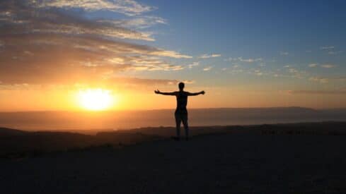 Person with arms outstretched overlooks sunset with orange sky and distant landscape.