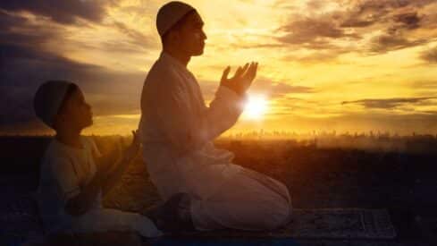 Man and child in white garments praying at sunset with city skyline in background.