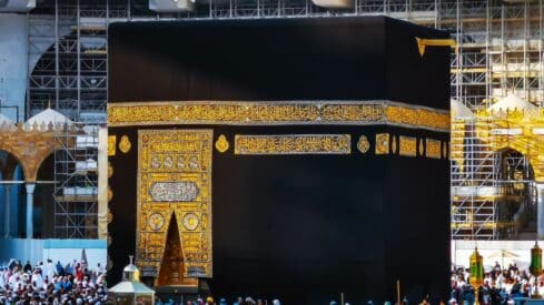 The Kaaba with black cloth and gold embroidery, surrounded by people and scaffolding.