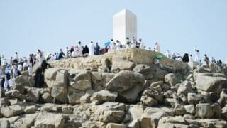 People in traditional attire on a rocky hill with a tall Arafat , white monument; umbrellas for shade.