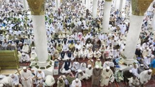 A large group gathered in a mosque with white columns, wearing light-colored garments.