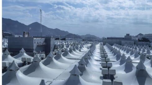 Vast area with white tents in rows, mountainous backdrop, partly cloudy sky, buildings interspersed.