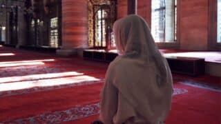 Person in white headscarf on red carpet in ornate building with sunlight patterns on the floor.