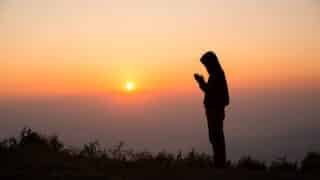 Person in hooded jacket silhouetted against sunset, sun low on horizon, with low vegetation.