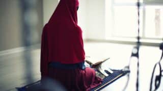 Person in red hijab kneeling on prayer mat with open book, natural light streaming in.