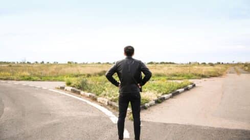 Person in black jacket at rural forked road, facing open fields and clear sky.