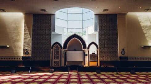 Interior of mosque with arched mihrab, patterned walls, skylight, carpet, and wall clock.