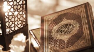 Decorative Quran cover with intricate patterns and a lit ornamental metal lantern beside it.