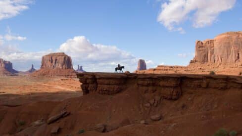 Lone rider on a horse crosses desert rock amidst towering mesas under a partly cloudy sky.