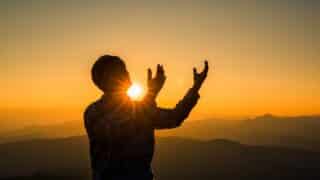 Silhouetted person with raised arms against a sunrise or sunset over mountains and clear sky.