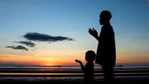 Adult and child silhouettes stand on beach at sunset, hands raised in reflection.