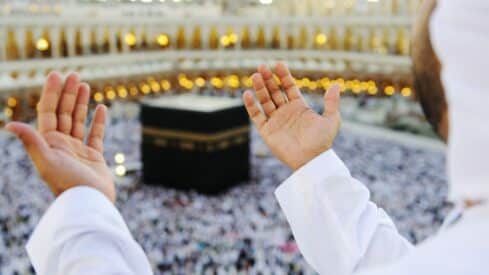 Person with hands raised in prayer facing the Kaaba, surrounded by a crowd and lit mosque.