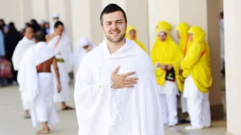 Man in white Ihram at Hajj with others; women in yellow attire in background.