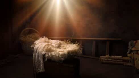 Dimly lit rustic room with a straw-lined manger, basket, and wooden ladder. Warm light above.