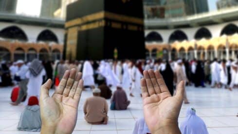 Hands raised in prayer at the Kaaba, Mecca; crowd in traditional attire surrounds it.