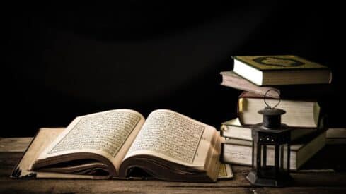 Open book with Arabic text on wooden table, surrounded by stacked books and lantern.